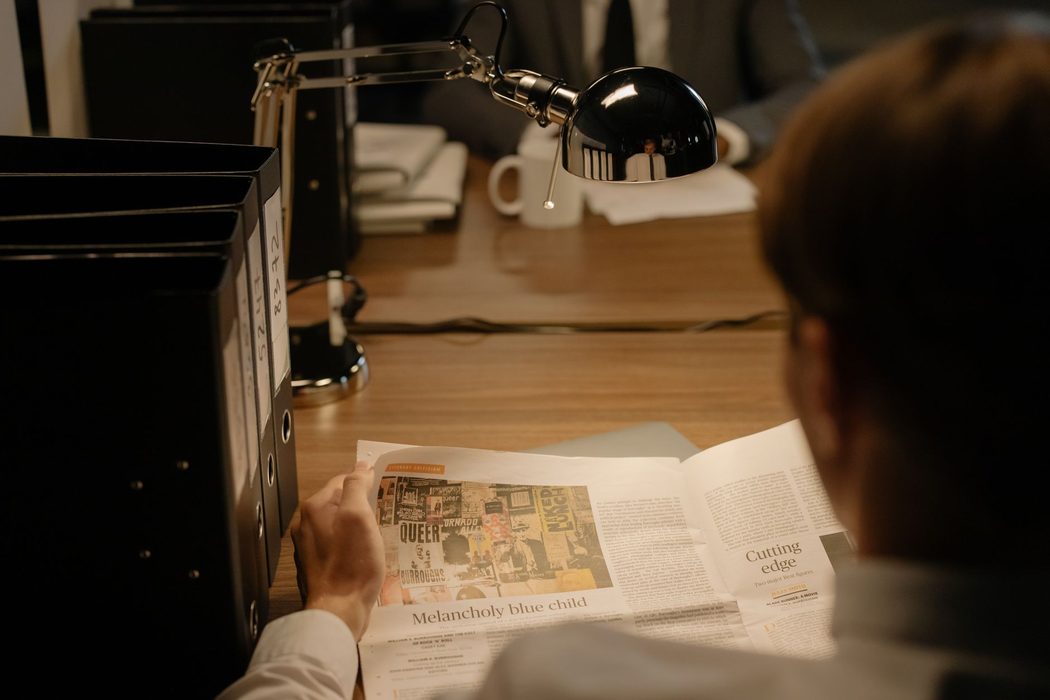 A person reading a printed newspaper at a desk covered with notes
