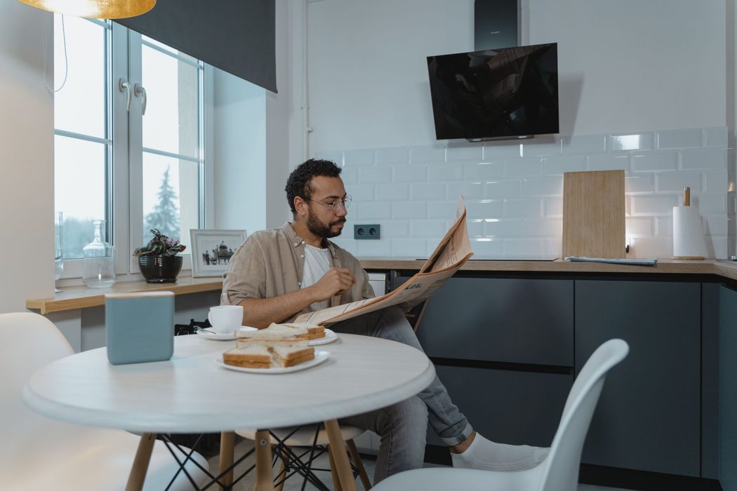 A person reading a newspaper at a kitchen table with a cup of coffee
