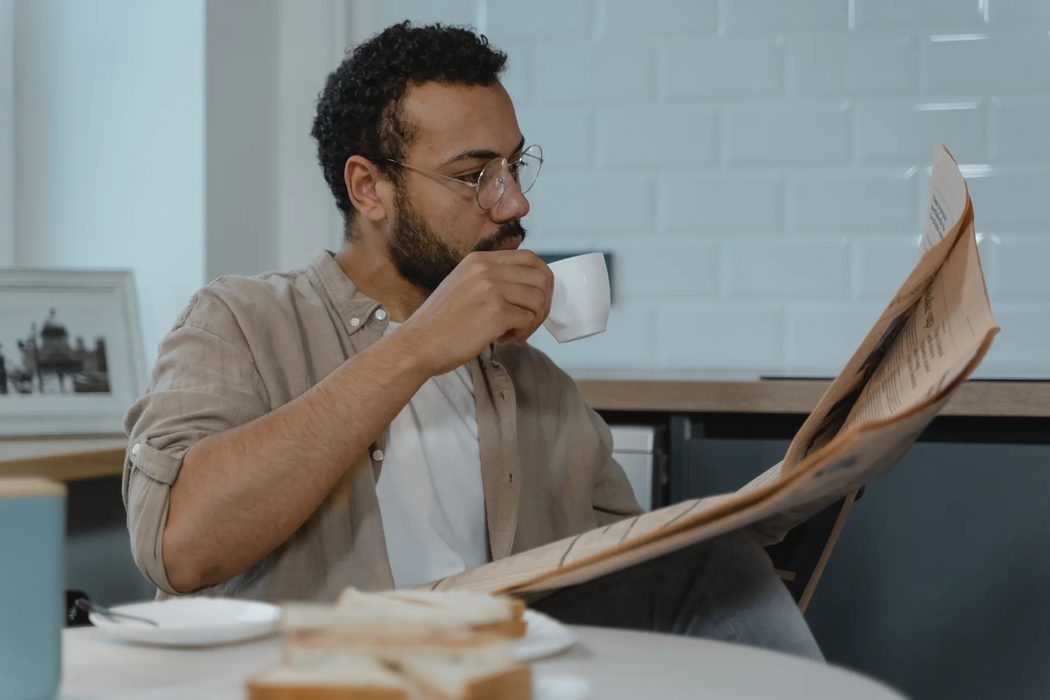 A reader with a printed newspaper and coffee at a kitchen table